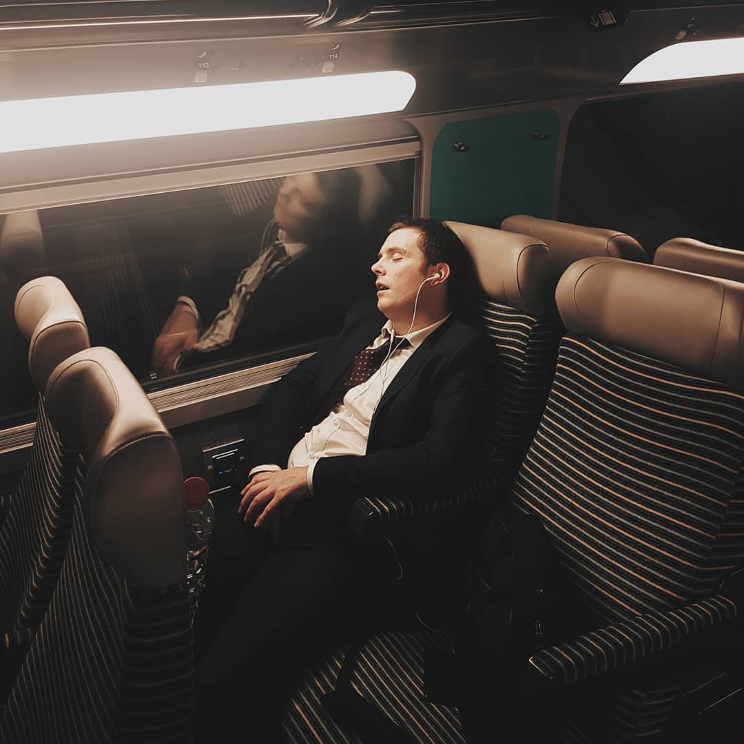 Adult male in a suit sleeping on a train with headphones in, illuminated by overhead light and reflection visible in window.