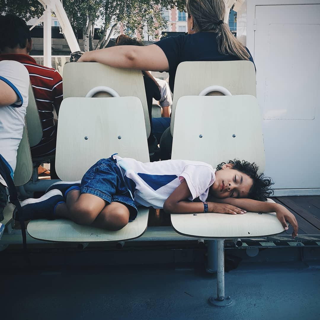 Young child asleep on white chair outdoors, wearing shorts and T-shirt, with other people in background. Travel and family scene.