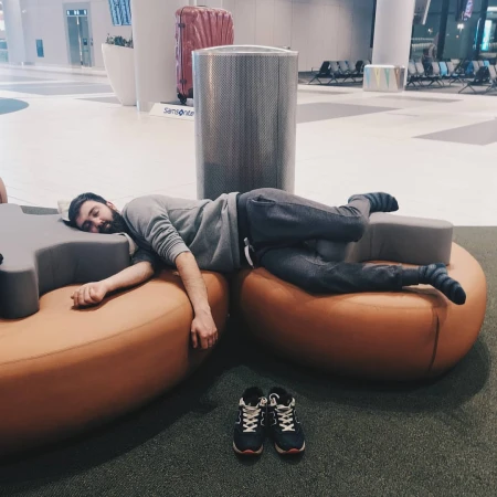 Man sleeping on lounge chairs at airport with sneakers nearby. Modern airport interior with gray, orange, and white tones.