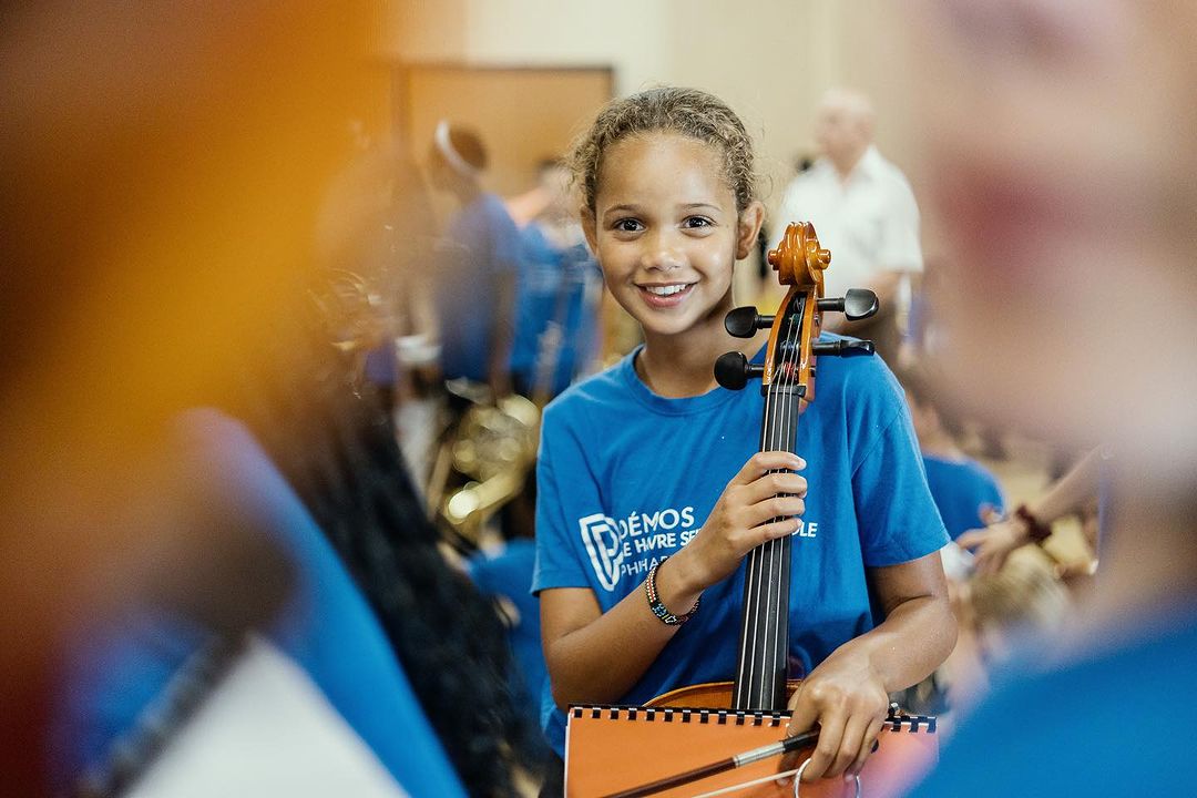 Smiling girl in blue shirt holding a cello in an indoor music lesson setting with other young musicians in the background.
