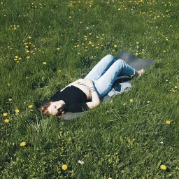 A woman lying on a yoga mat in a green field filled with scattered yellow wildflowers under bright sunlight.
