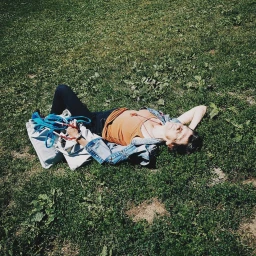 Person lying down in grass with arms behind head, wearing a denim jacket, orange top, and dark trousers next to white bags.