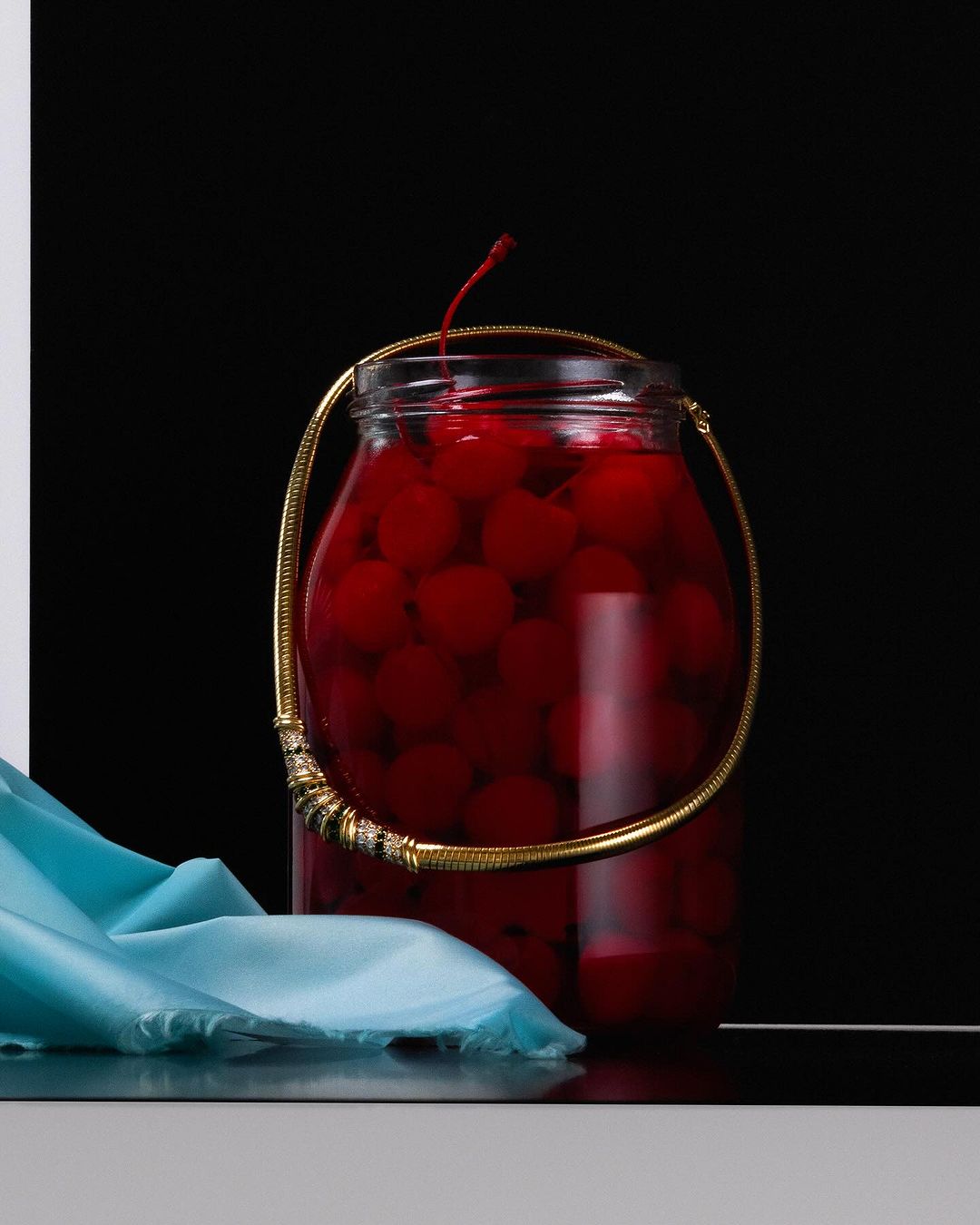 A glass jar is filled with bright red cherries, accessorized with a gold necklace and a draped turquoise fabric, set against black and white.