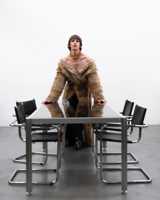 Adult male in fur coat standing behind glass table with chrome chairs in a minimalist white studio setting with concrete flooring.