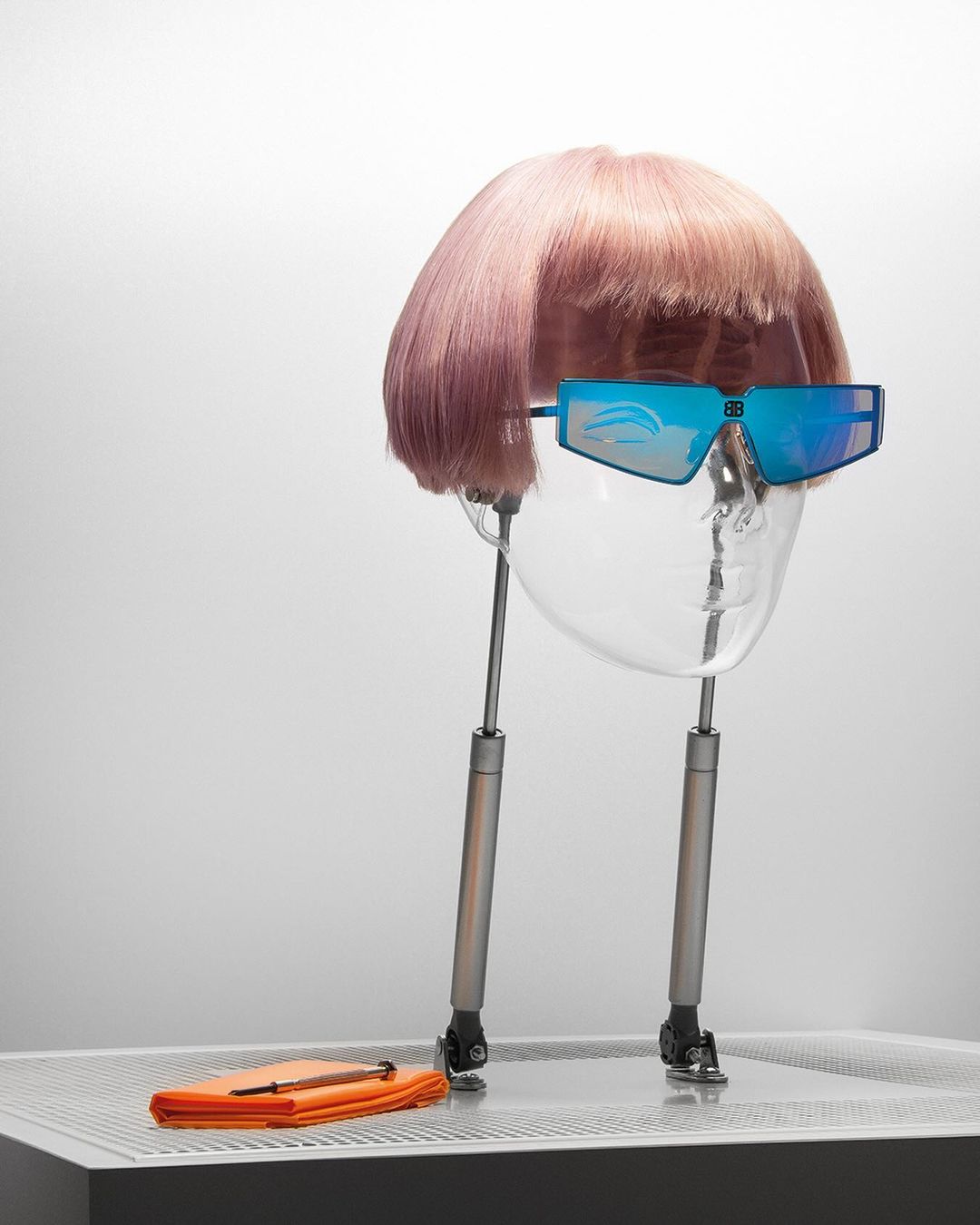 A mannequin head with pink hair and futuristic sunglasses sits on a metal platform with an orange cloth, isolated against white background.
