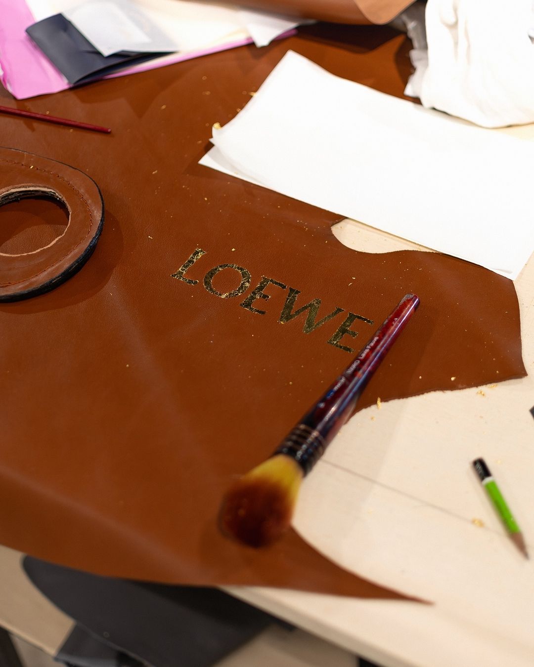 Loewe logo on brown leather, with a makeup brush and paper scraps on a white surface, creating a textured studio scene.