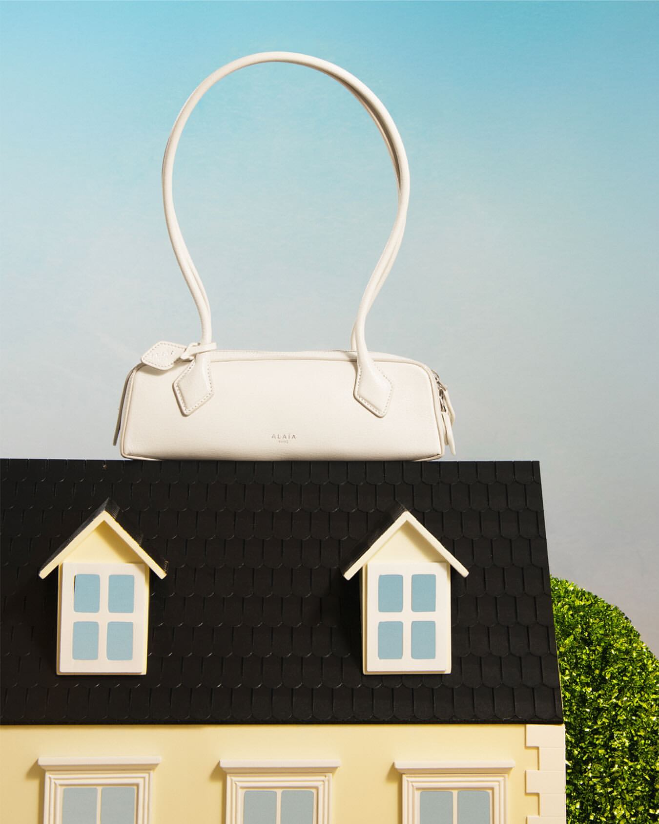 White leather purse perched on top of a toy house with two windows and a green bush in a product photography style shot.