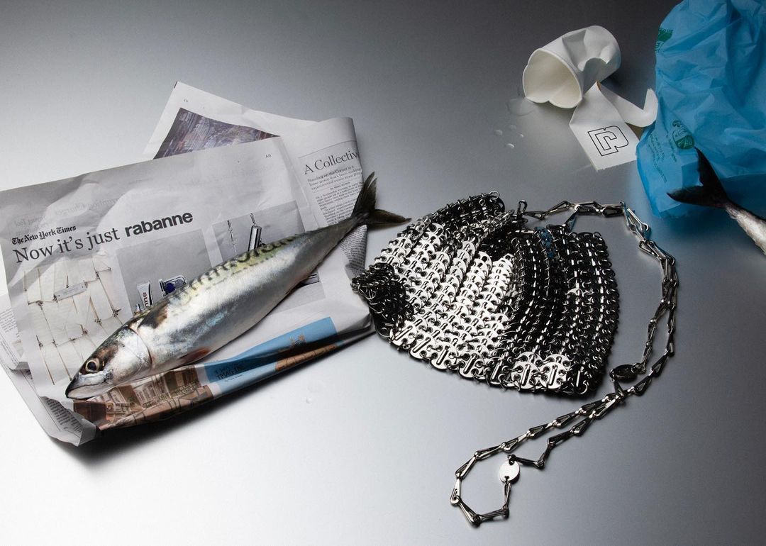 Still life featuring a raw mackerel on a newspaper next to a metal mesh purse with crumpled paper cups and a blue plastic bag.