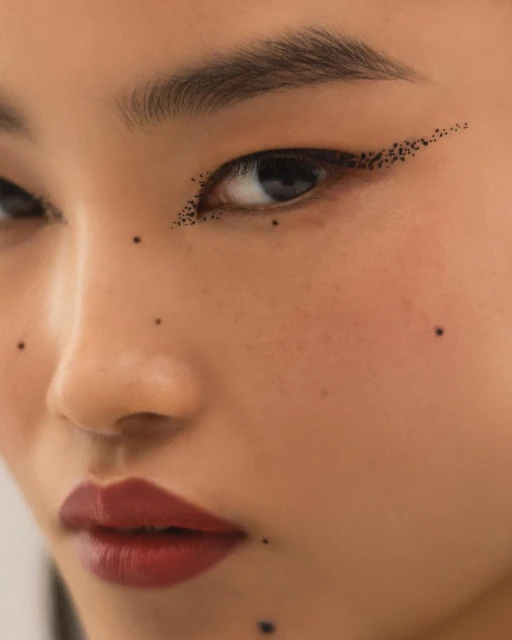 Close-up portrait of an Asian woman with dark red lipstick and black dotted eyeliner in a beauty editorial shot. Minimalist look.