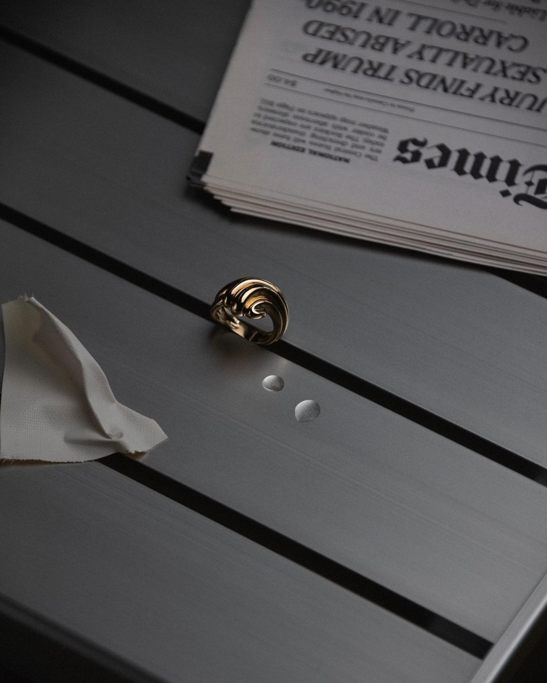 Close-up studio shot featuring a gold ring, two water droplets, a white cloth, and a newspaper on a gray, striped table surface.