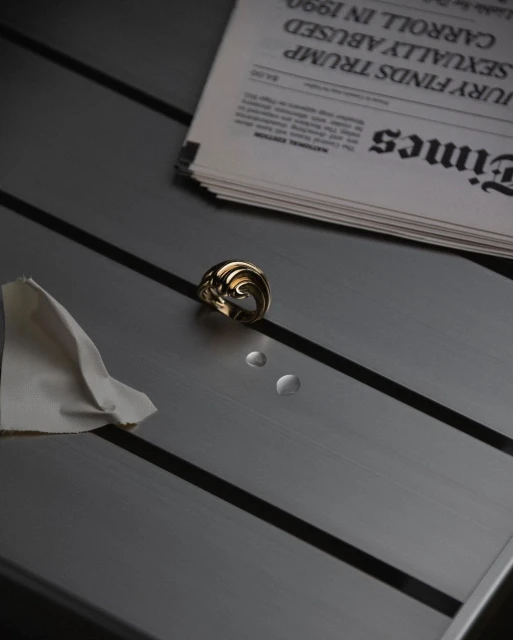 Close-up studio shot featuring a gold ring, two water droplets, a white cloth, and a newspaper on a gray, striped table surface.