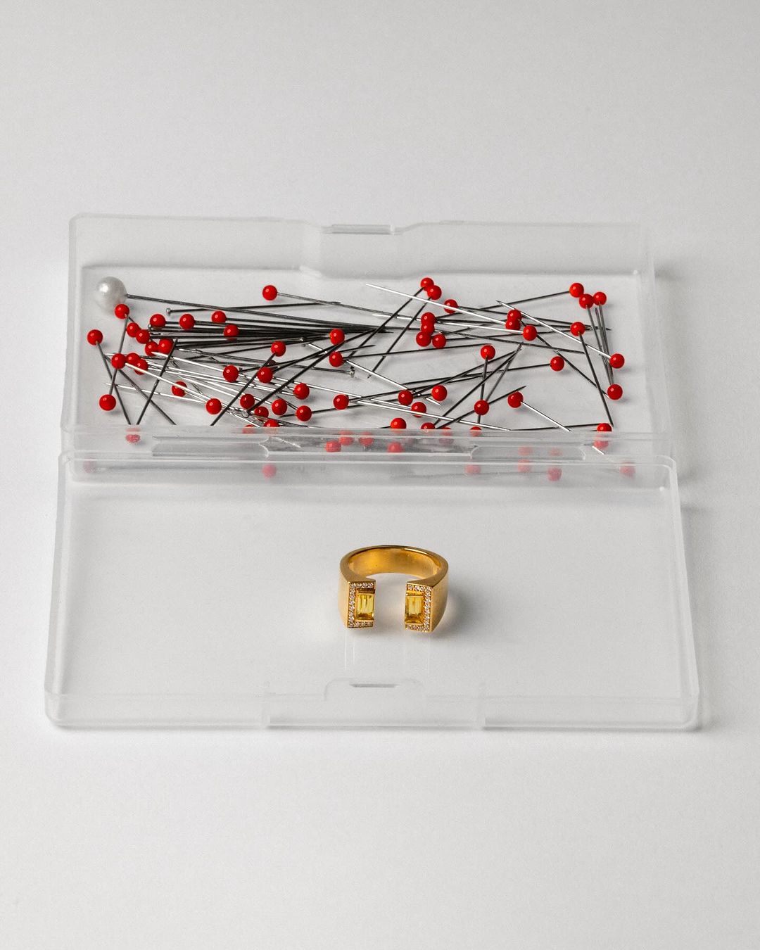 Gold colored modern ring with yellow gemstones inside a clear plastic container with sewing pins on a white background.