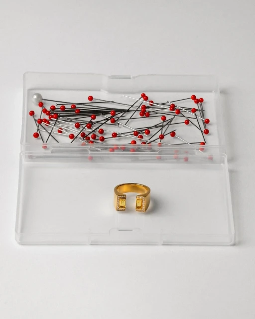 Gold colored modern ring with yellow gemstones inside a clear plastic container with sewing pins on a white background.