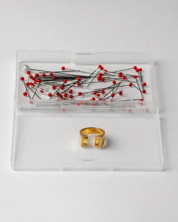 Gold colored modern ring with yellow gemstones inside a clear plastic container with sewing pins on a white background.