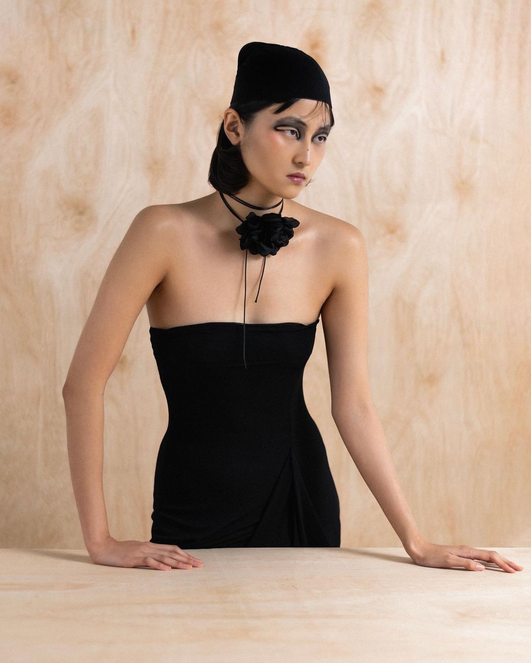 An Asian woman with a beanie poses in a strapless dress with a large flower choker against a neutral backdrop in a studio portrait.