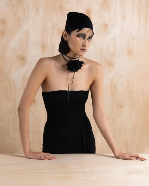 An Asian woman with a beanie poses in a strapless dress with a large flower choker against a neutral backdrop in a studio portrait.