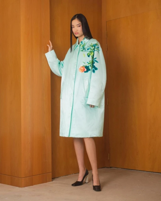 An adult female model poses in a light-blue floral-painted coat against a neutral yellow-beige background, wearing black high heel shoes.