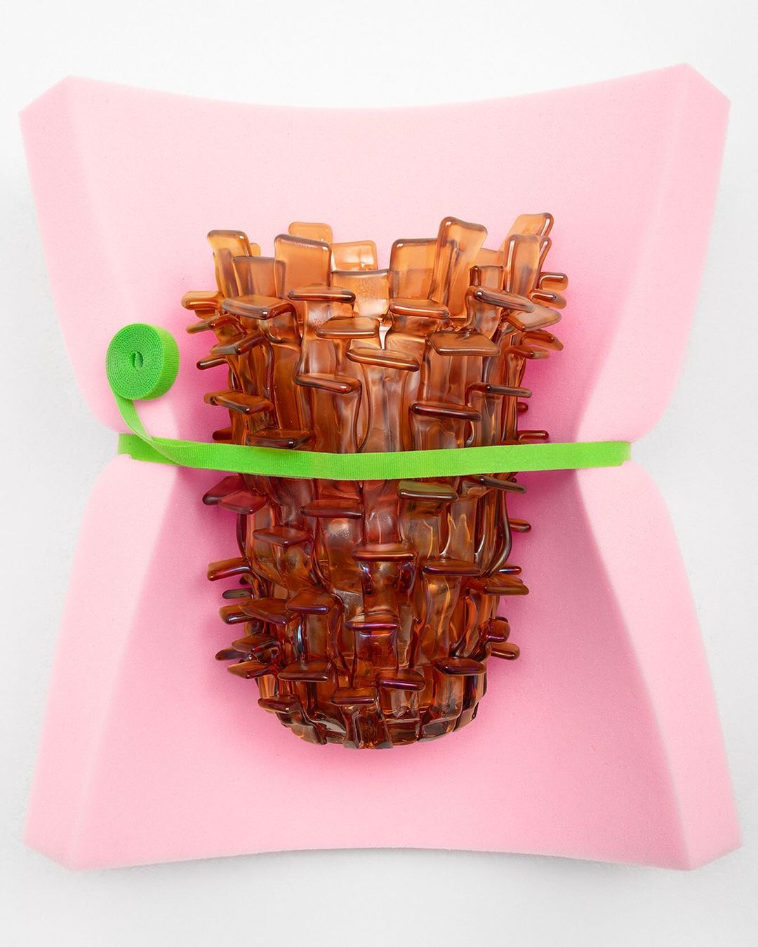 Artistic still life featuring a textured amber glass vase secured with a vibrant green band against a soft pink support. 