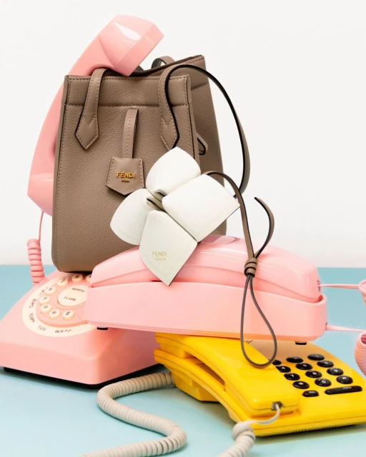 Beige leather Fendi handbag atop stacked retro pink and yellow rotary phones, studio lit against a bright cyan background.