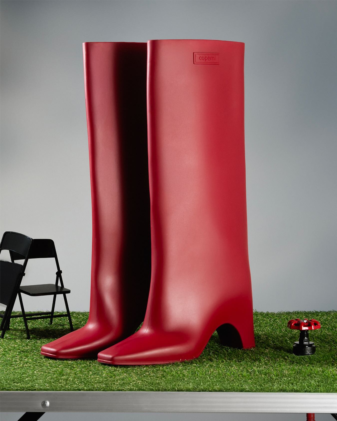 Striking fashion still life: Two tall, shiny, red rubber boots with heels, set on artificial turf in a studio environment. Minimalist and bold.