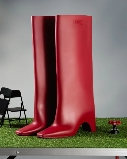 Striking fashion still life: Two tall, shiny, red rubber boots with heels, set on artificial turf in a studio environment. Minimalist and bold.
