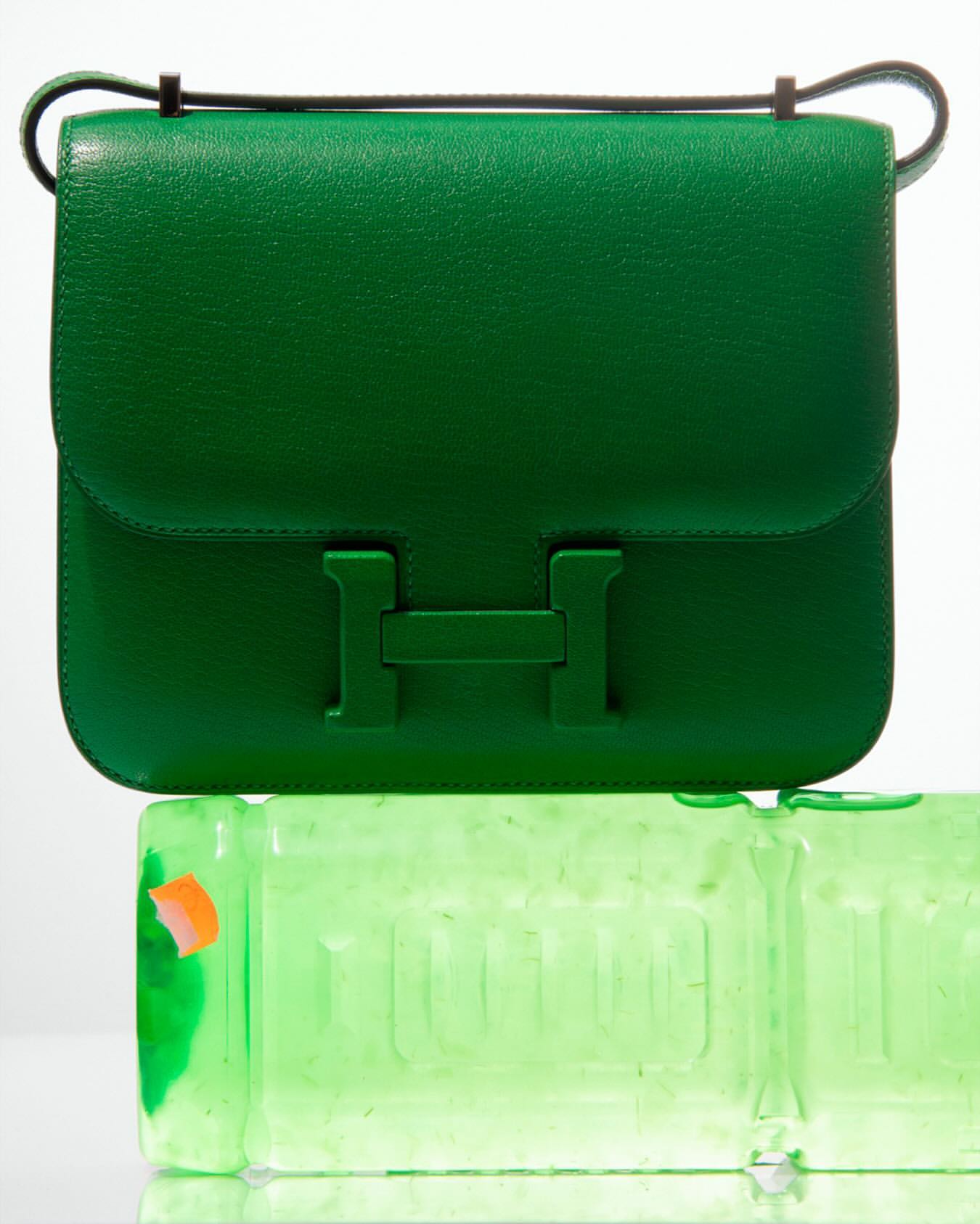 A green leather purse sits atop a clear, partially filled plastic container, set against a bright white background in a studio-lit scene.