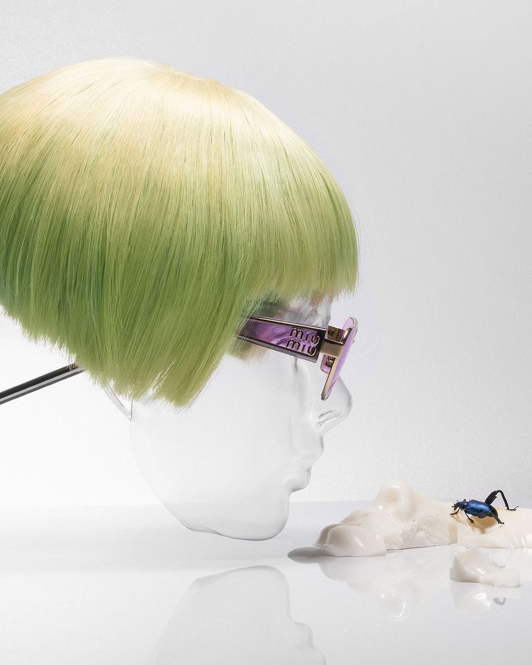 Fashion shot of a clear plastic mannequin head with a green and blonde wig wearing pink-framed sunglasses next to a blue beetle.