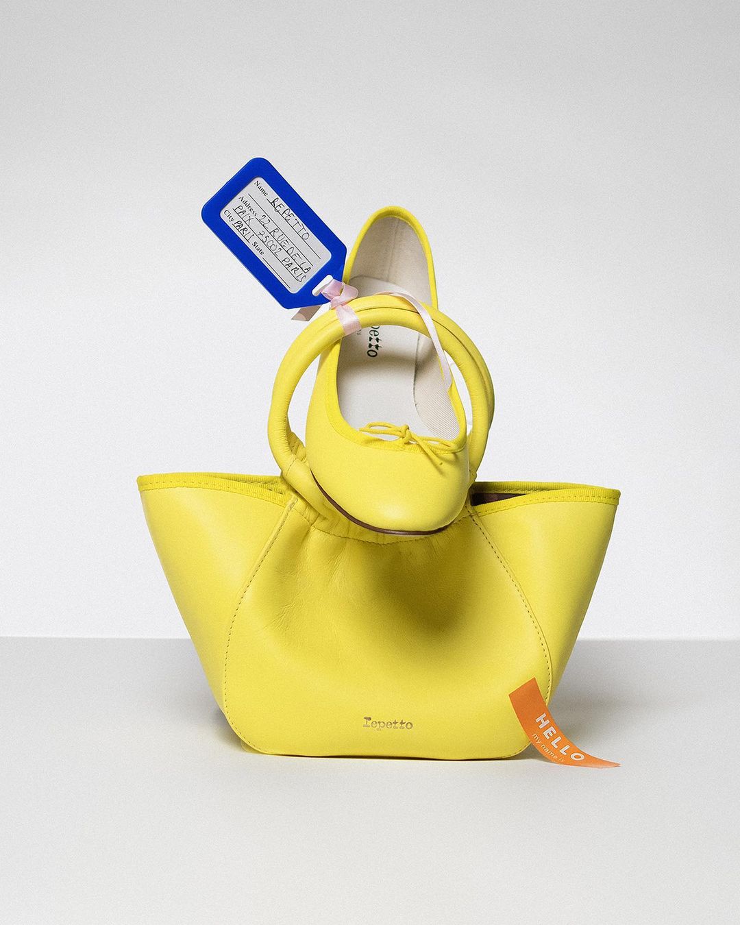 Bright yellow Petit Bateau leather handbag with ballet shoe placed on top and a blue tag, photographed in a photo studio.