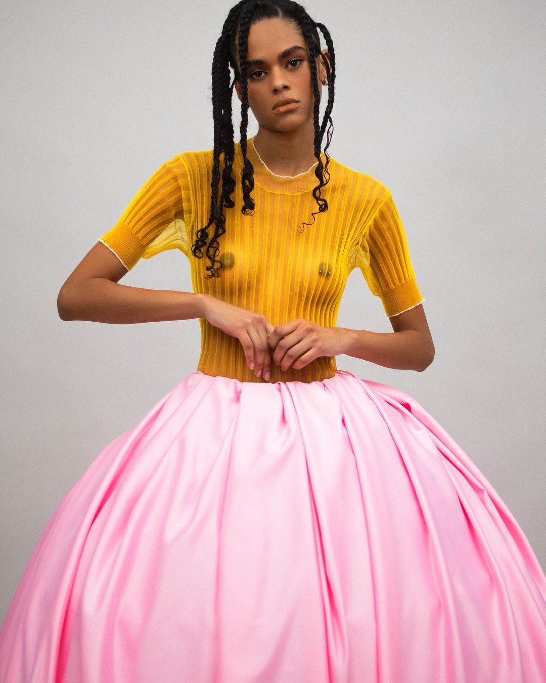 Woman with braided hair wears a yellow top and large pink skirt in a studio shot. Fashion and portrait photography in a neutral setting.
