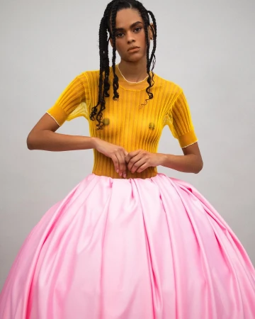 Woman with braided hair wears a yellow top and large pink skirt in a studio shot. Fashion and portrait photography in a neutral setting.