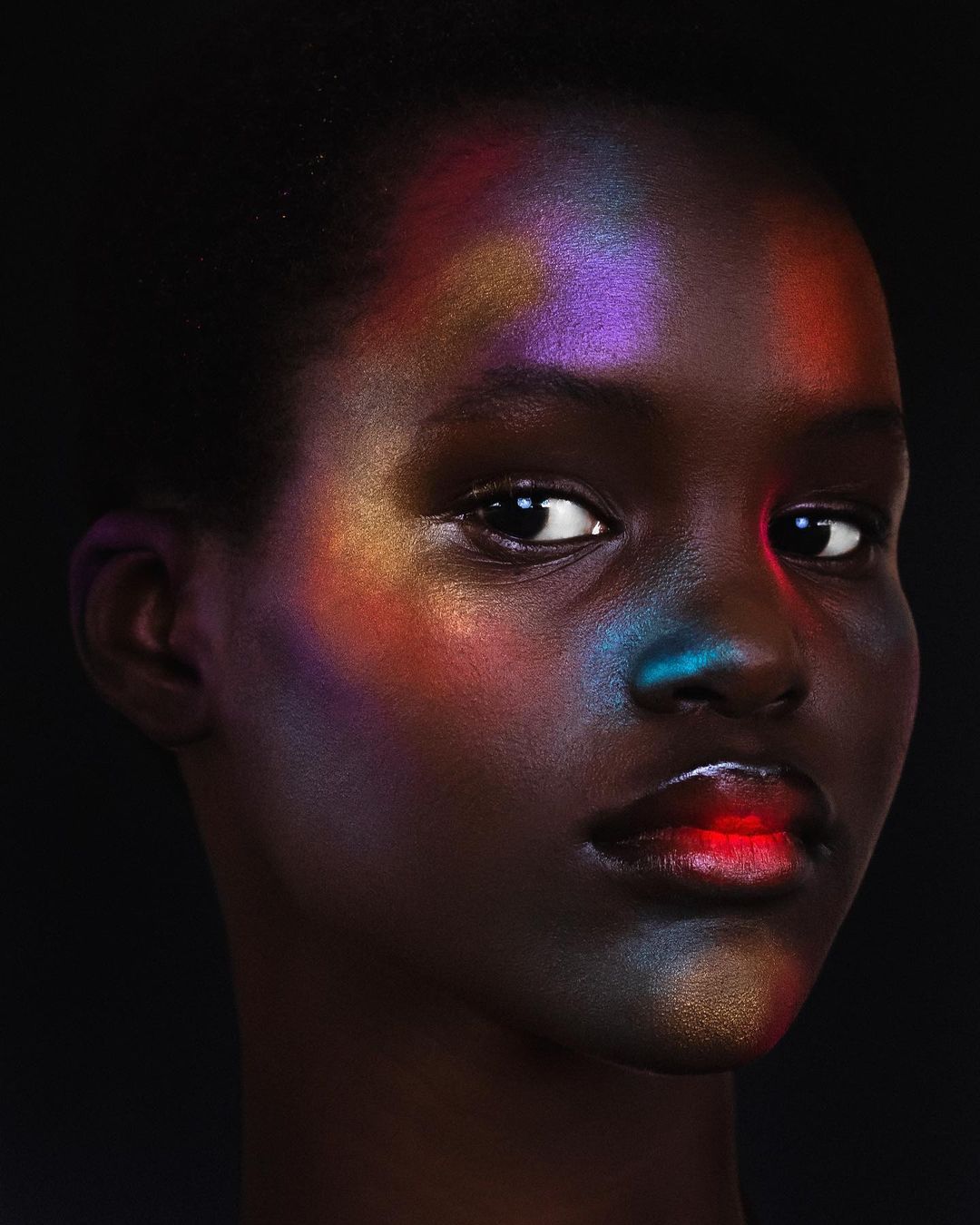 Close-up portrait of a Black woman with colorful iridescent light projections on her face against a black background.