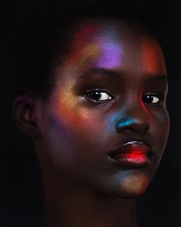 Close-up portrait of a Black woman with colorful iridescent light projections on her face against a black background.