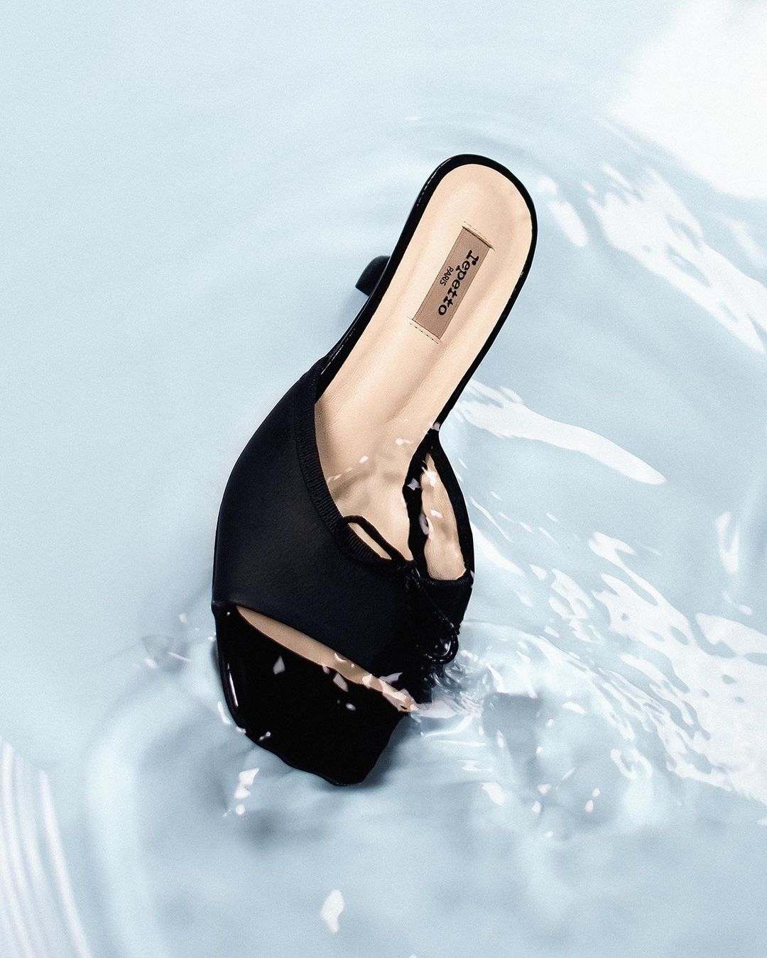 Elegant black mule shoe by Repetto Paris sits in light blue water. Close-up product shot with visible water textures and studio lighting.