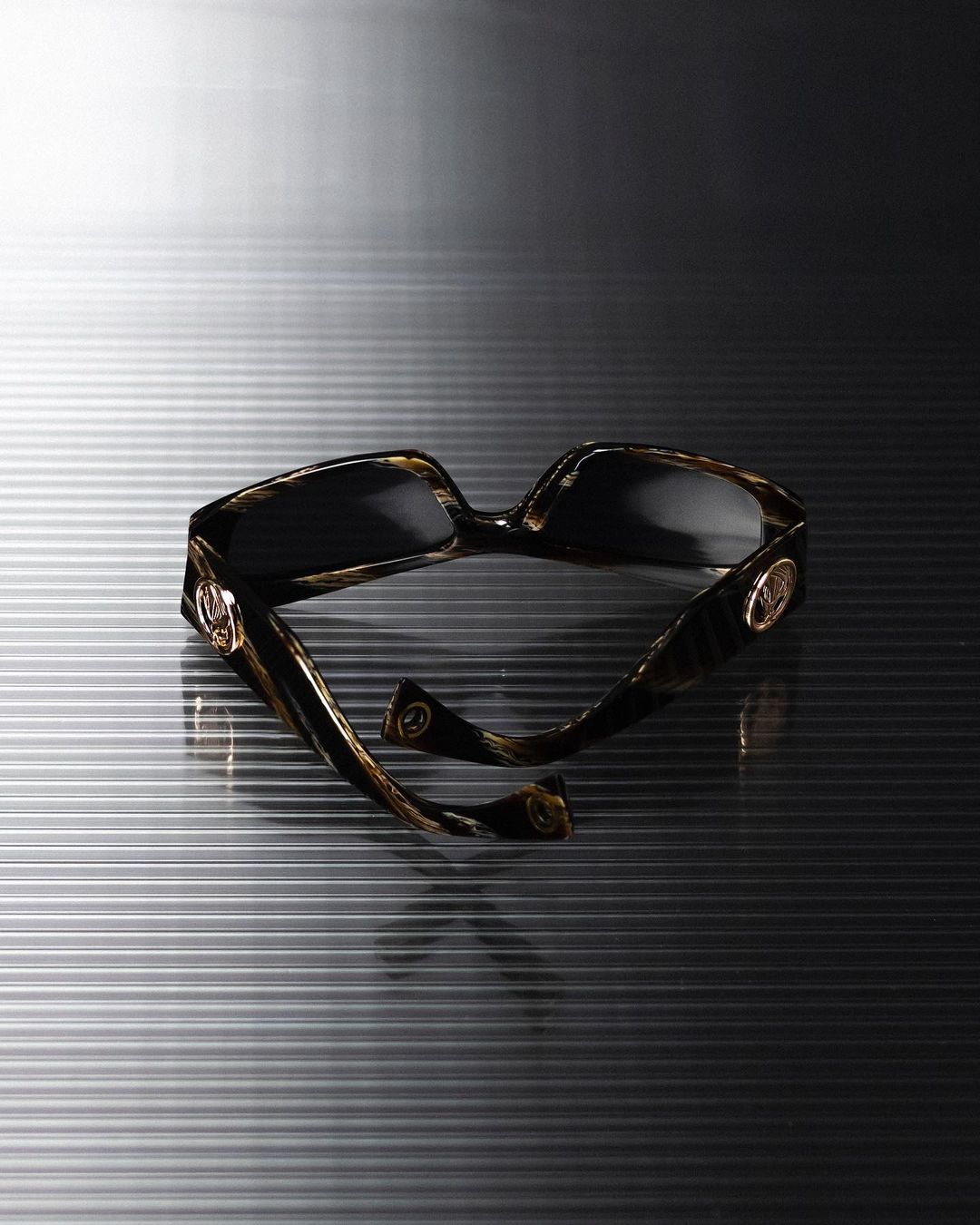 Tortoiseshell sunglasses with gold circular hardware resting on a ribbed grey metallic surface.