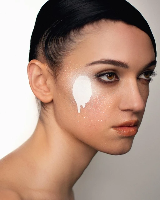 Close-up studio shot of a young adult woman with fair skin, slicked-back hair, and a white cream-like substance dripping on her face.