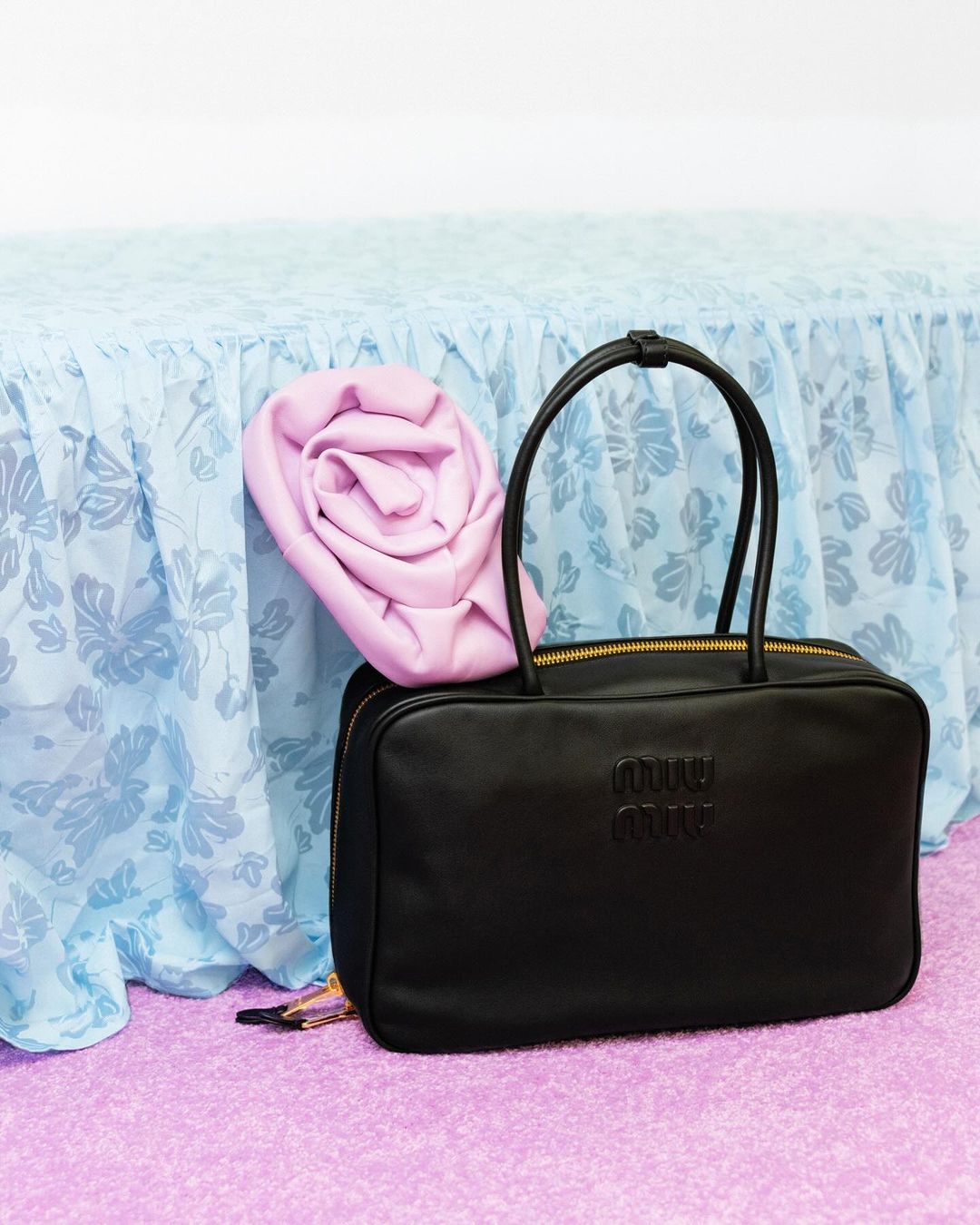 Black leather Miu Miu handbag with logo, gold hardware and a light pink garment, against a floral patterned backdrop.