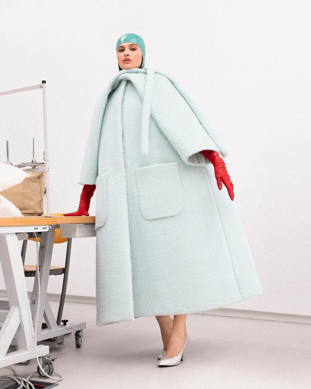 Woman in oversized light blue coat, matching hat, and red gloves poses next to sewing table in minimalist studio, looking at the camera.