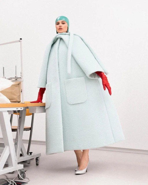 Woman in oversized light blue coat, matching hat, and red gloves poses next to sewing table in minimalist studio, looking at the camera.