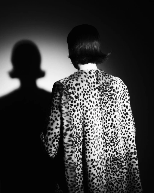 Back view of a woman with short dark hair wearing a black and white spotted coat standing in front of a wall casting a shadow.