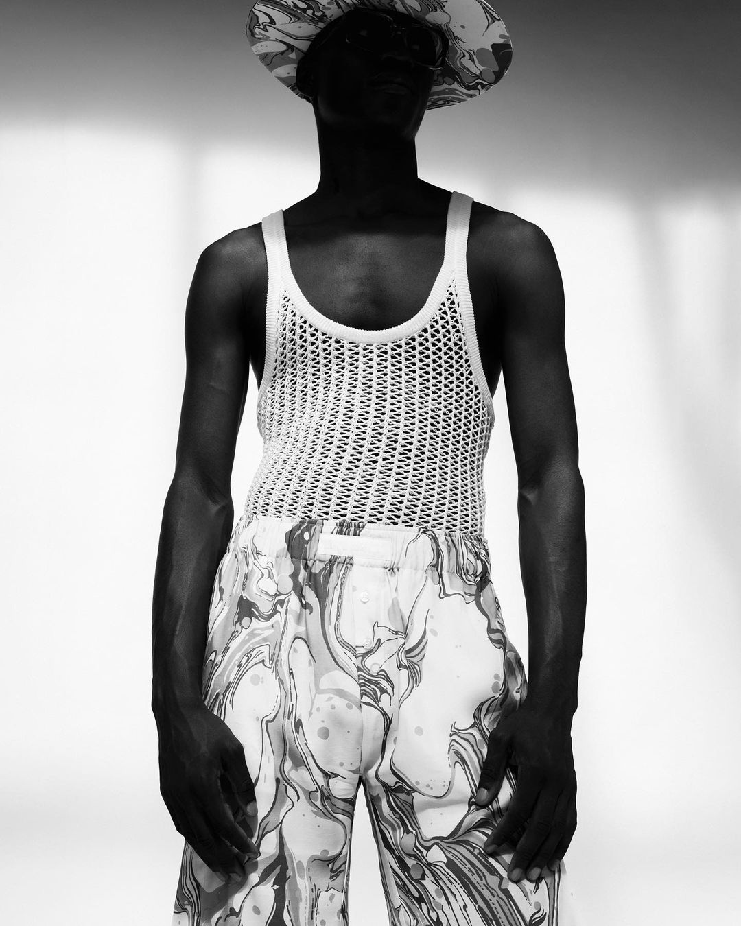 An adult male wearing a patterned hat and pants poses in front of a window. The subject is also wearing a white, knitted tank top.