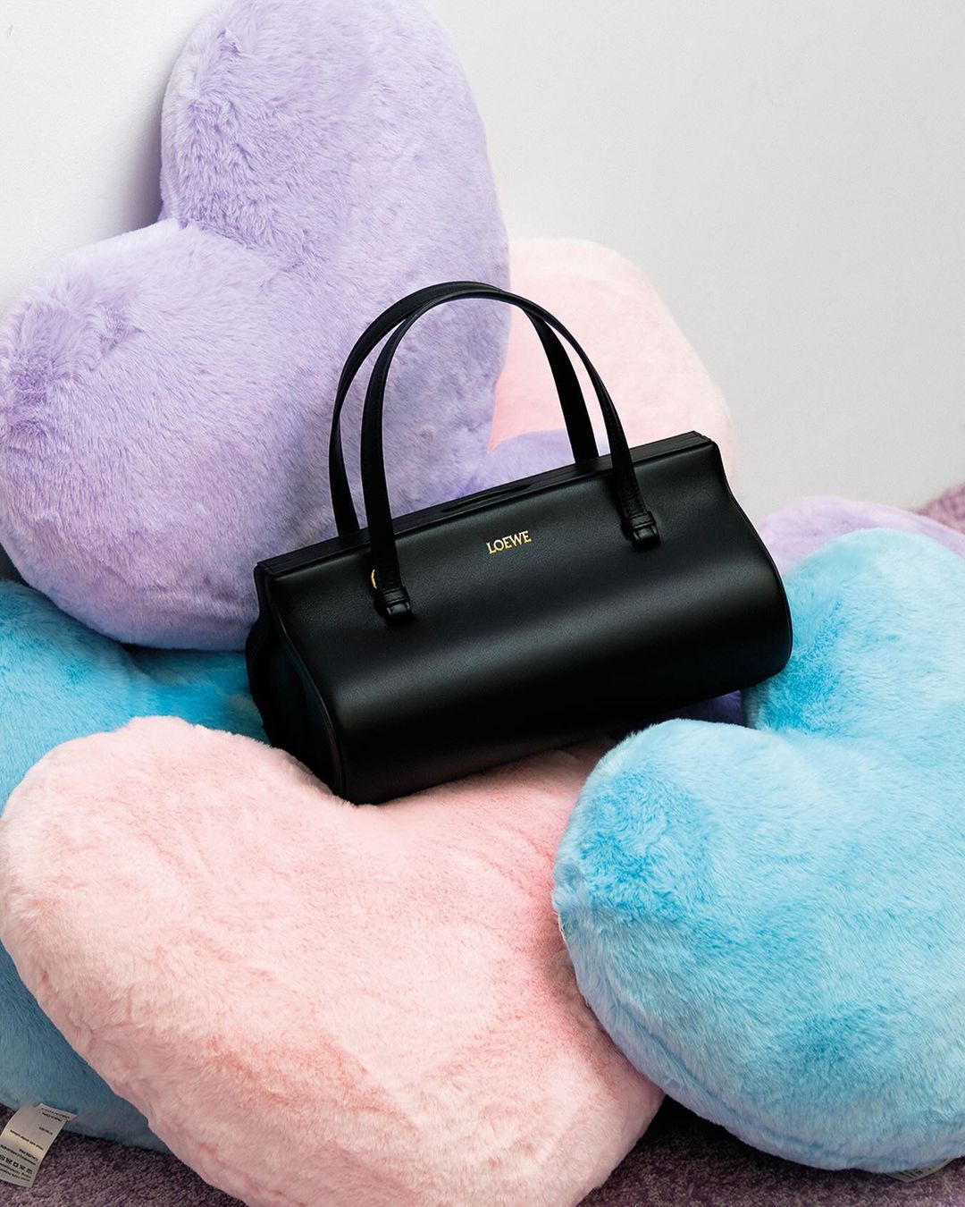 A black Loewe handbag sits on a pile of pastel heart-shaped pillows in a soft, inviting studio setting.