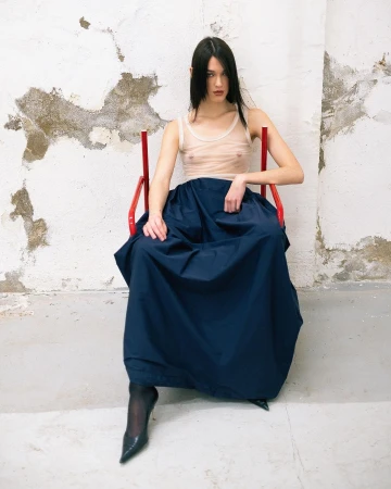 A slender woman with long dark hair sits on a red chair in a photo studio, wearing a translucent top and a large navy skirt.