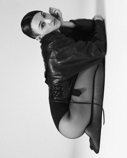 Black-and-white portrait of a woman in a black leather jacket, black pantyhose, and dark bodysuit, with a sleeked-back hairstyle.