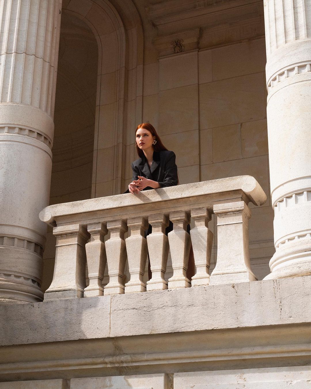 Elegant woman with auburn hair in a black blazer posing on a stone balcony in front of columns in a stone building.