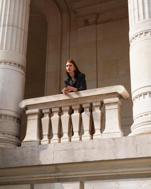 Elegant woman with auburn hair in a black blazer posing on a stone balcony in front of columns in a stone building.