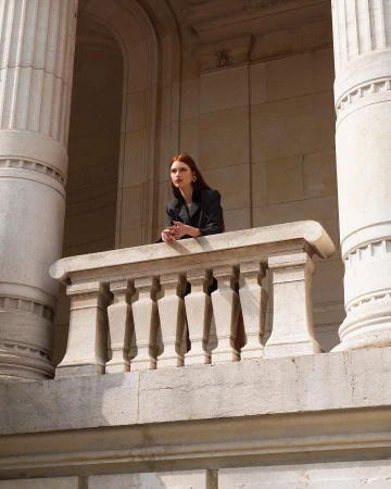 Elegant woman with auburn hair in a black blazer posing on a stone balcony in front of columns in a stone building.
