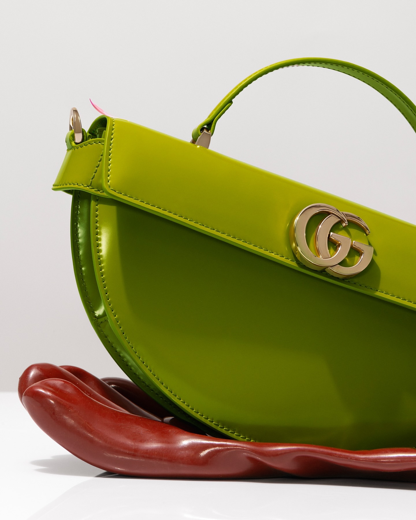 Studio shot of a vibrant green Gucci handbag with gold logo hardware, displayed on a glossy, curved, hand-shaped, red-brown prop.