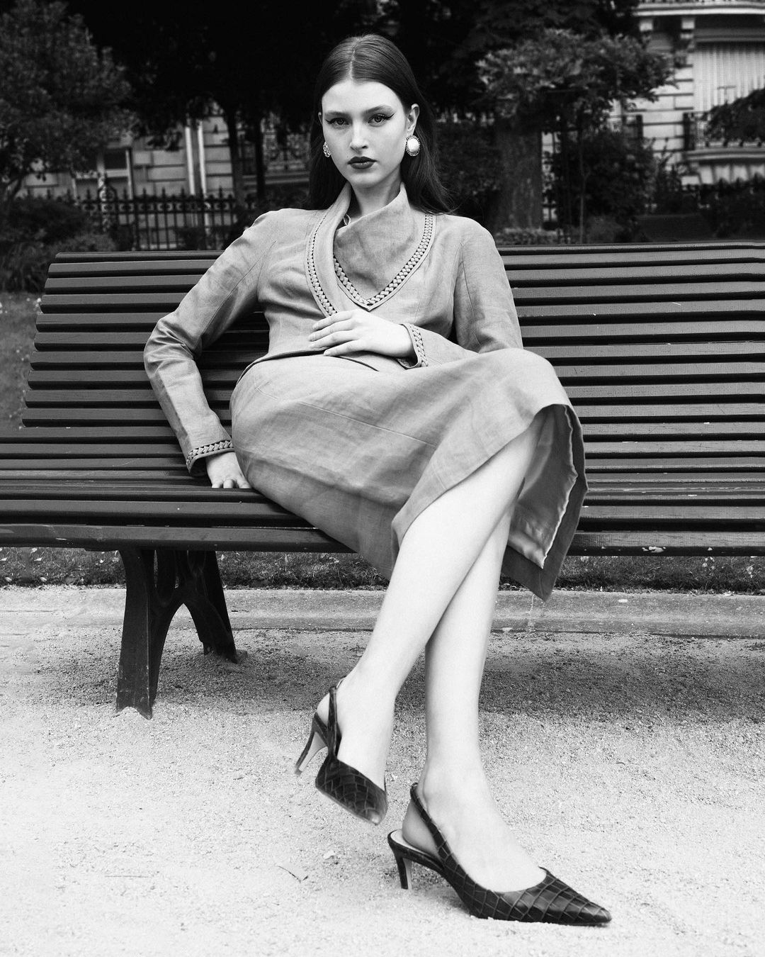 Elegant woman in dress and heels sits on park bench in black and white, gazing at camera with soft, natural light illuminating her.