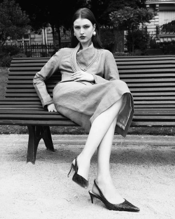 Elegant woman in dress and heels sits on park bench in black and white, gazing at camera with soft, natural light illuminating her.