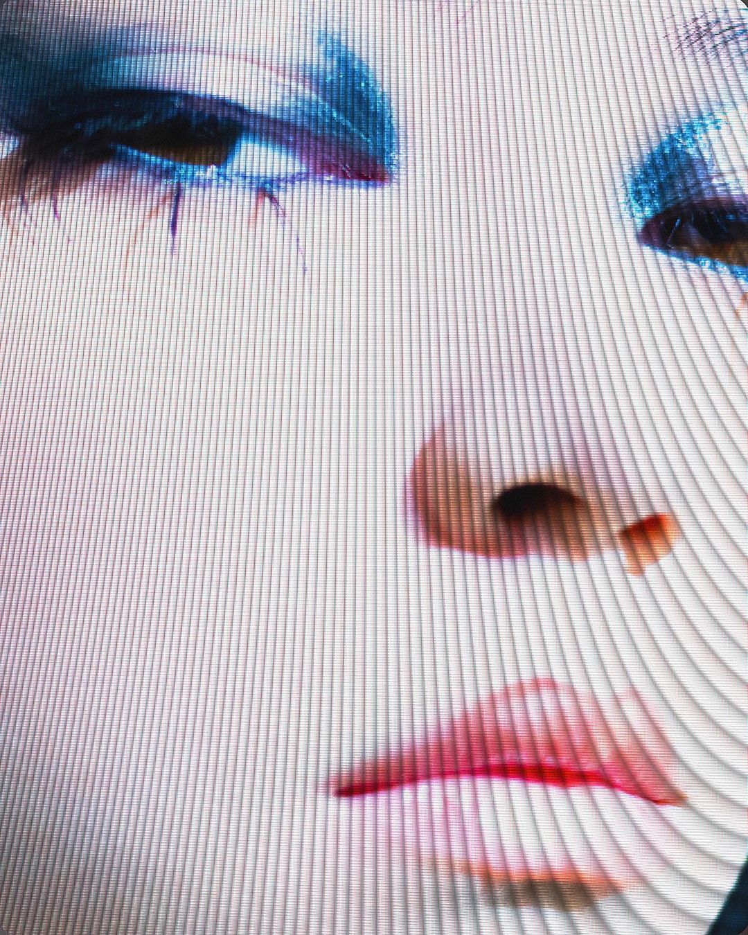Close-up of a woman's face with blue eyeshadow, visible on a screen with a linear pattern overlay, showcasing the nose and red lips.